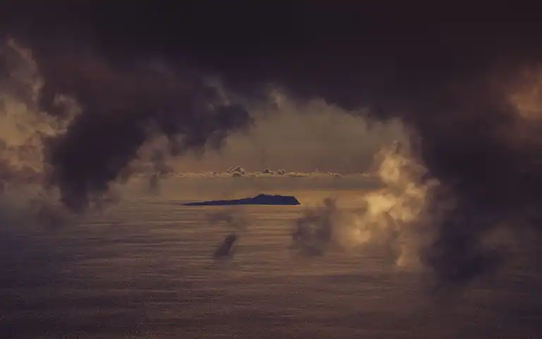 Storm clouds hang over a distant island and dark ocean beneath a band of cloud near the horizon.