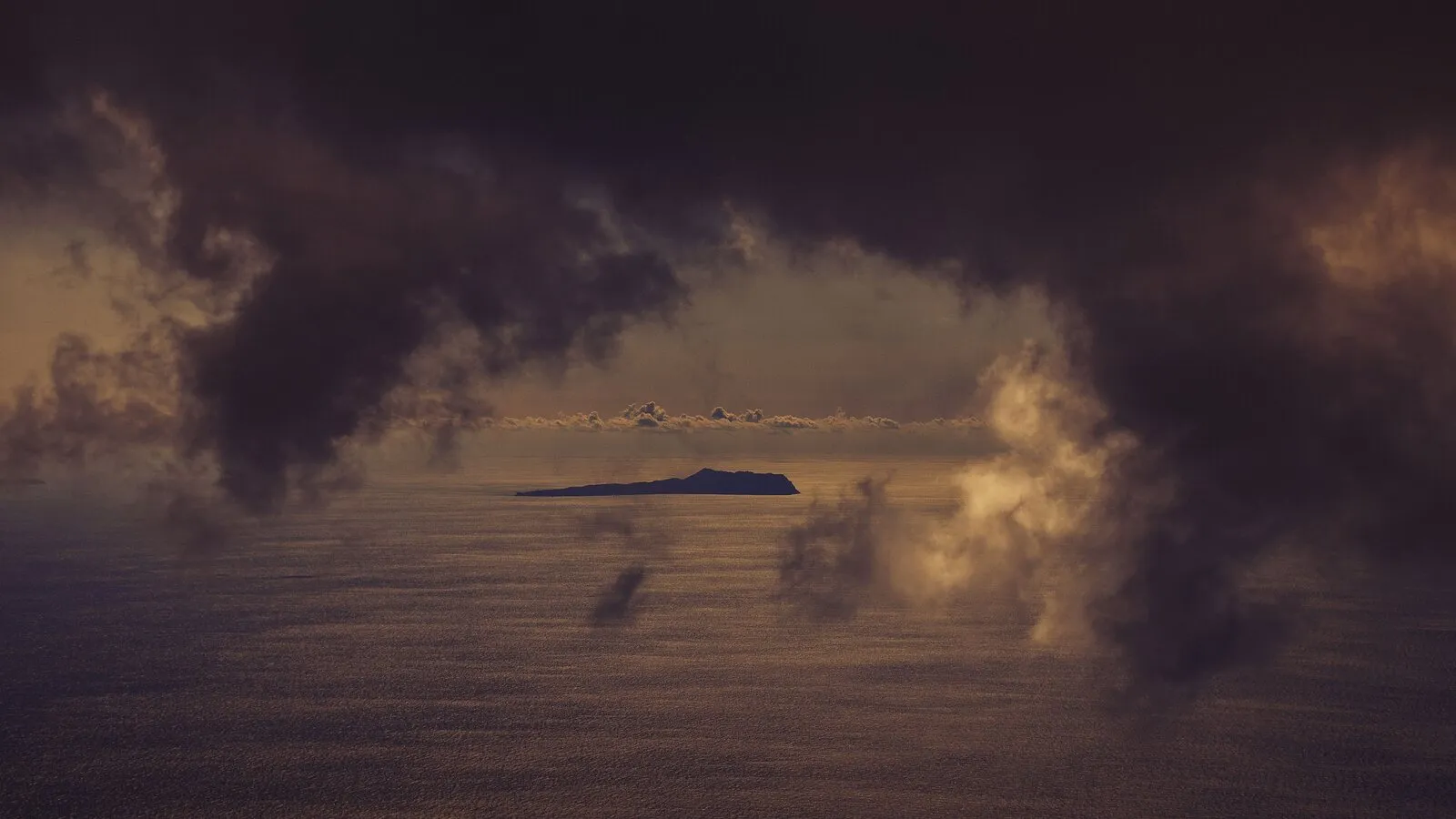 Storm clouds hang over a distant island and dark ocean beneath a band of cloud near the horizon.