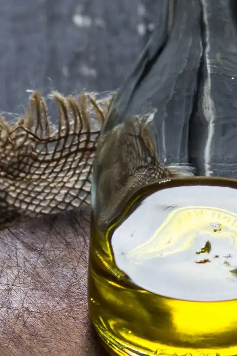 A clear glass bottle of golden oil on a wooden cutting board against a dark woven backdrop.