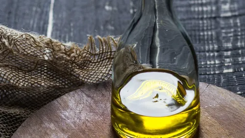 A clear glass bottle of golden oil on a wooden cutting board against a dark woven backdrop.
