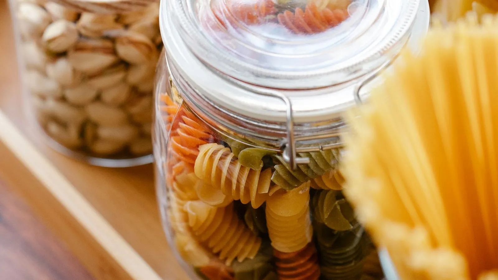 Glass jars containing dry spiral pasta and other pantry ingredients on a wooden tray.