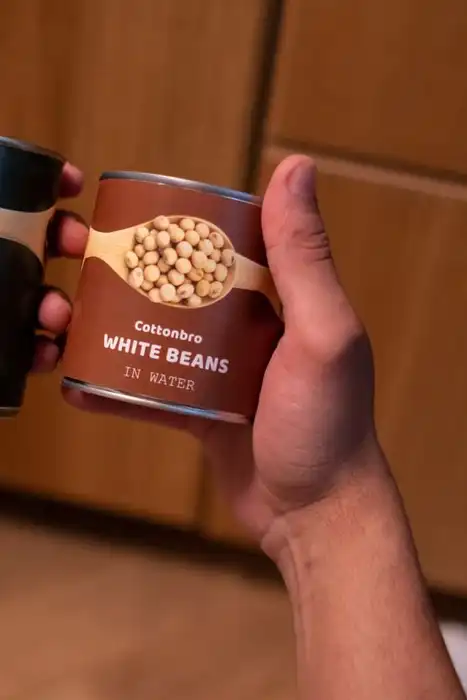 Hands holding two cans of beans in a kitchen.
