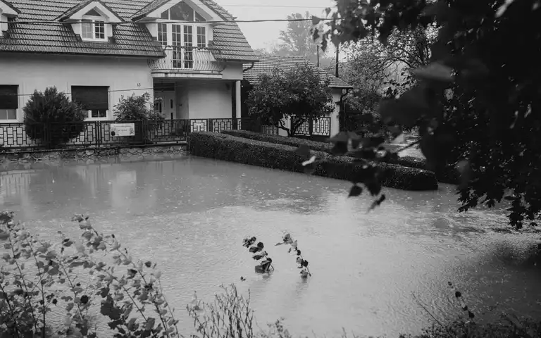 Rain falls on floodwater surrounding a house and garden in a black-and-white photograph.