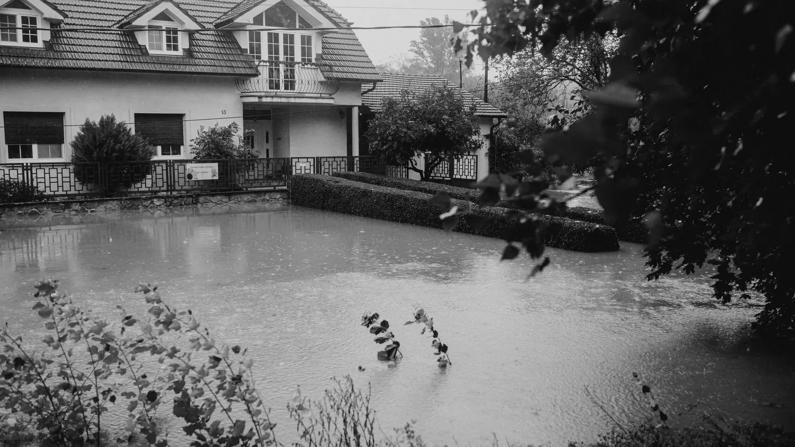 Rain falls on floodwater surrounding a house and garden in a black-and-white photograph.
