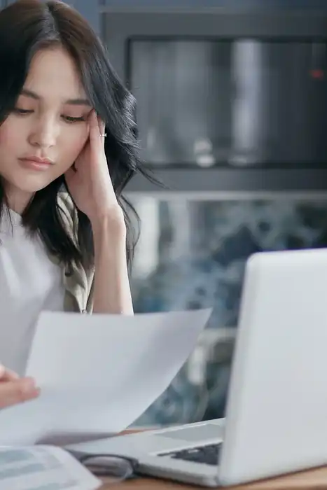 Woman reviewing a sheet of household paperwork beside a laptop at home