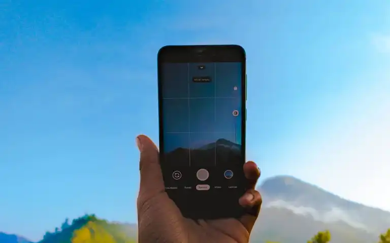 A hand holds a smartphone toward a mountain landscape.