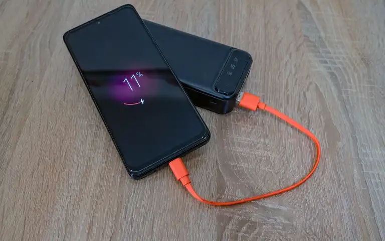 A smartphone connected to a portable power bank by an orange cable on a wooden table.