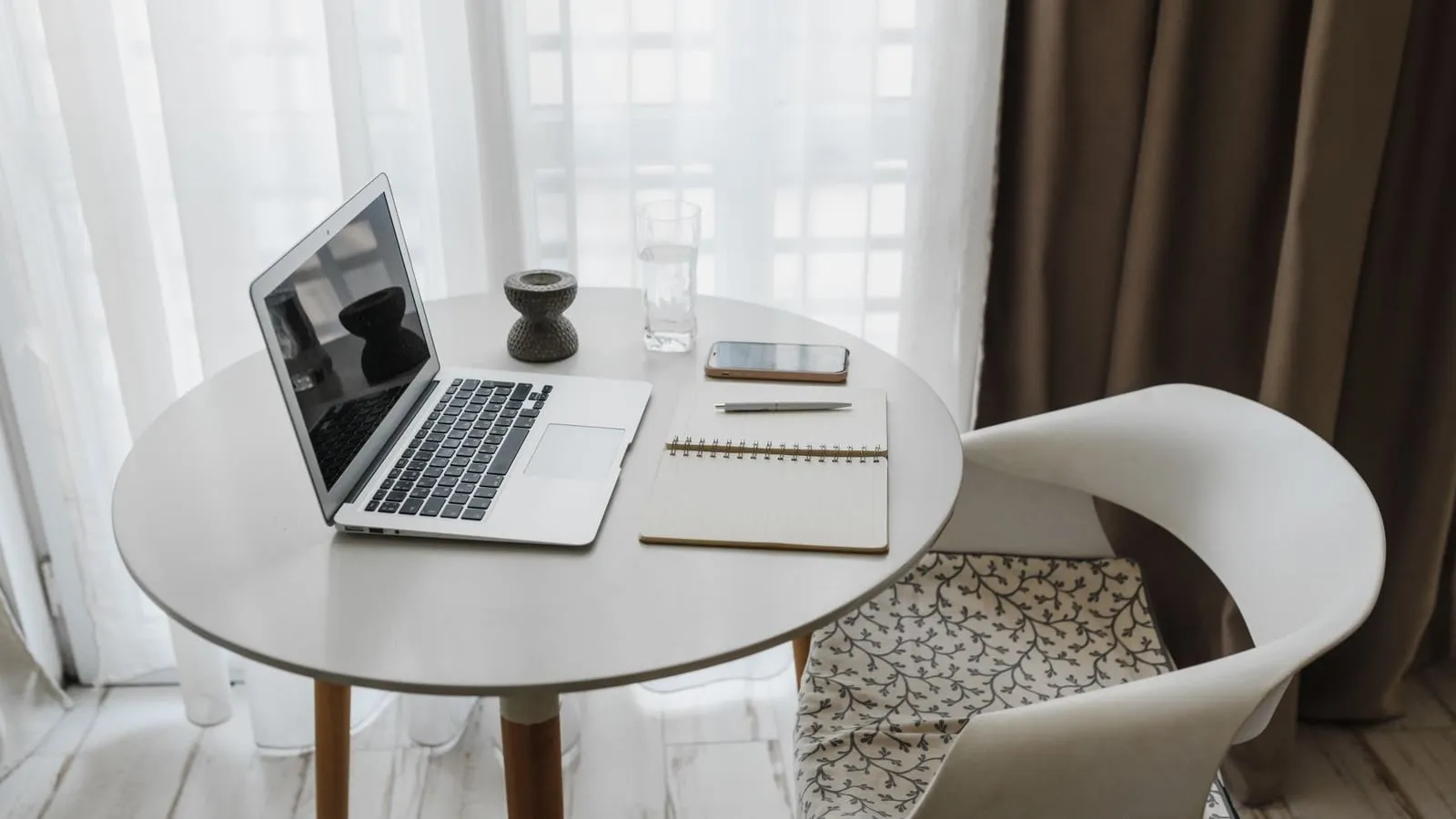 An open laptop, notebook and phone on a round table beside a bright window.
