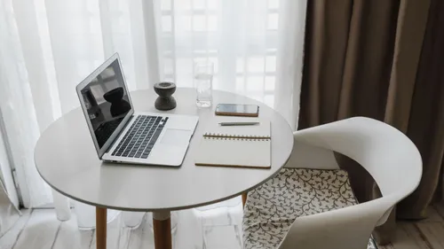 An open laptop, notebook and phone on a round table beside a bright window.