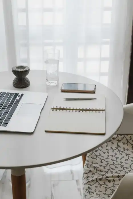 An open laptop, notebook and phone on a round table beside a bright window.