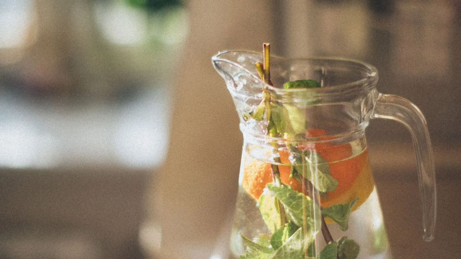 A clear pitcher of water with fruit and leaves stands on a table.