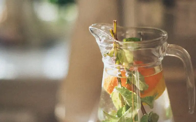 A clear pitcher of water with fruit and leaves stands on a table.