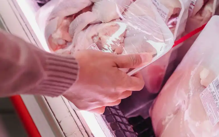 A shopper selects a tray of packaged raw chicken from a refrigerated case.