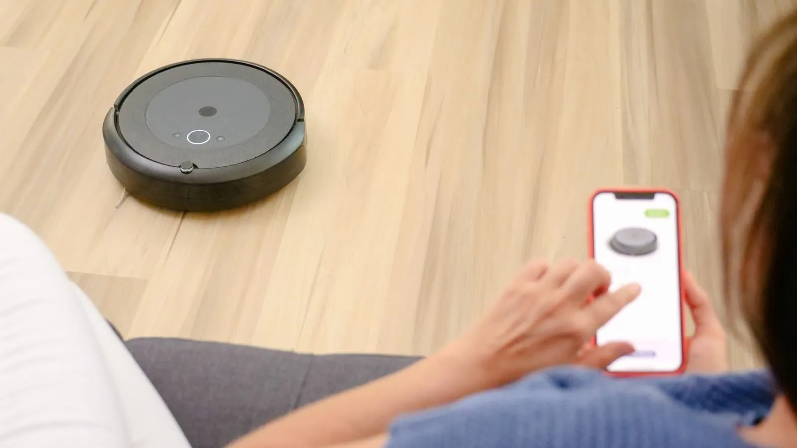 A person holds a smartphone near a robotic vacuum on a wood floor.