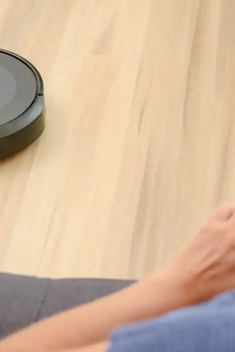 A person holds a smartphone near a robotic vacuum on a wood floor.