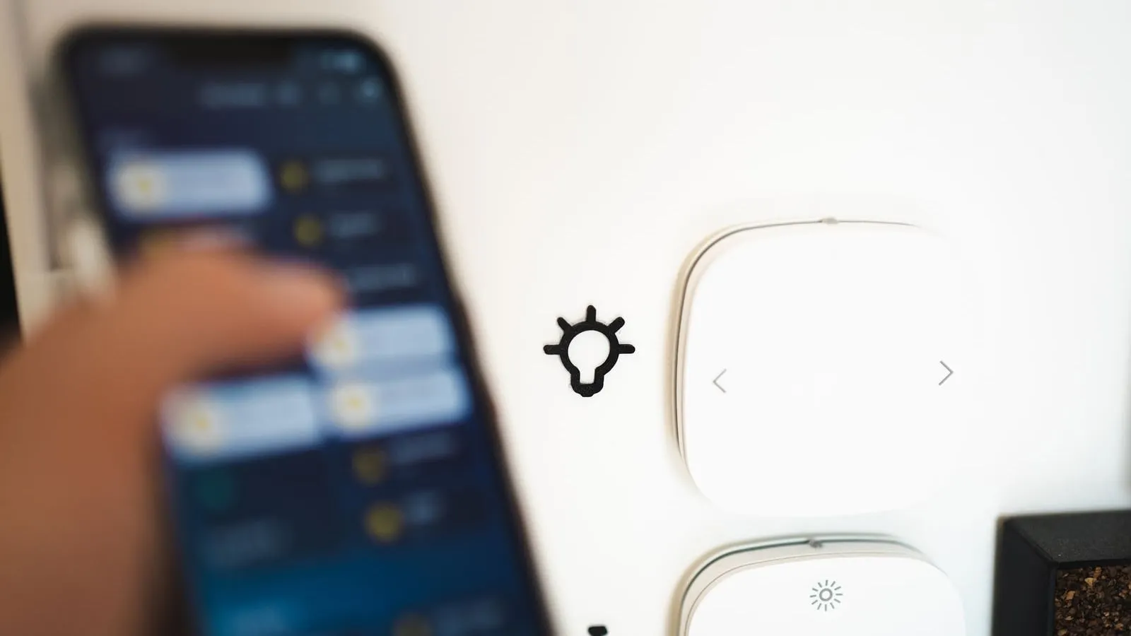 A hand holds a smartphone beside two smart-home wall controls.