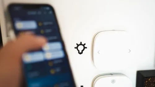 A hand holds a smartphone beside two smart-home wall controls.