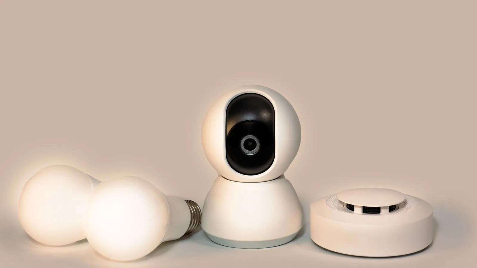 A smoke detector, camera, and two light bulbs are arranged on a plain surface.