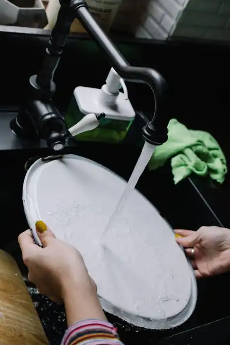 Hands wash a plate under running water in a kitchen sink.