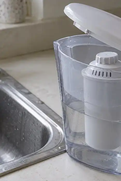 A hand lifting the lid of a generic filter pitcher beside a kitchen sink