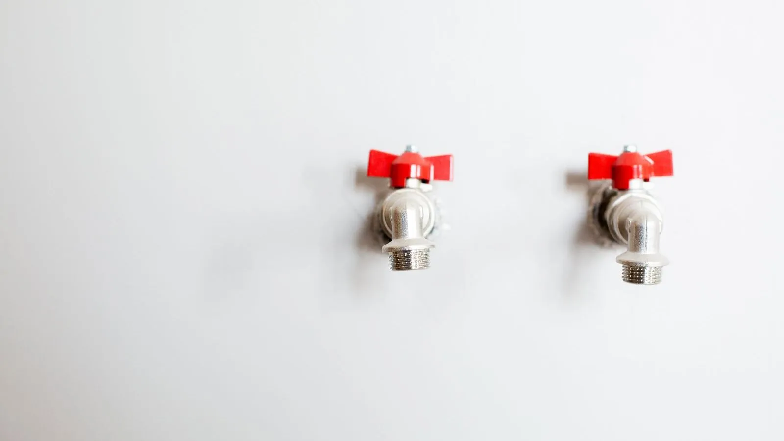 Two metal faucets with red handles are mounted on a white wall.