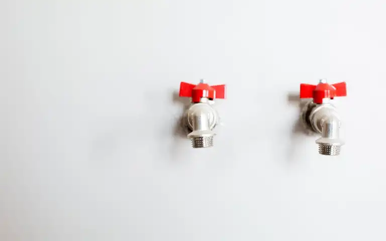 Two metal faucets with red handles are mounted on a white wall.